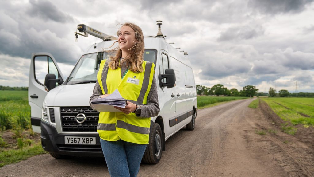 A woman stood in front of a white van in a high-vis jacket holding papers.