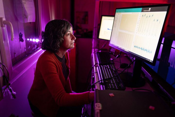 Woman sat in front of two computer screens looking at data. The room is dark with red, pink and purple lights on.