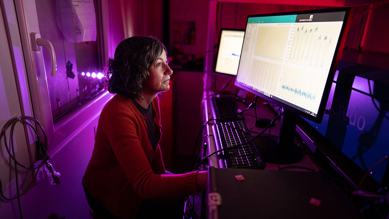 Woman sat in front of two computer screens looking at data. The room is dark with red, pink and purple lights on.
