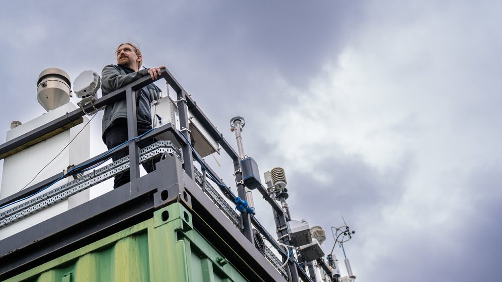 Person stands on rooftop holding a railing, looking skywards. There are scientific instruments attached along the railing.