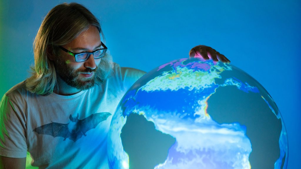 Person with long hair wearing glasses stands next to an illuminated globe, displaying a map of the world.