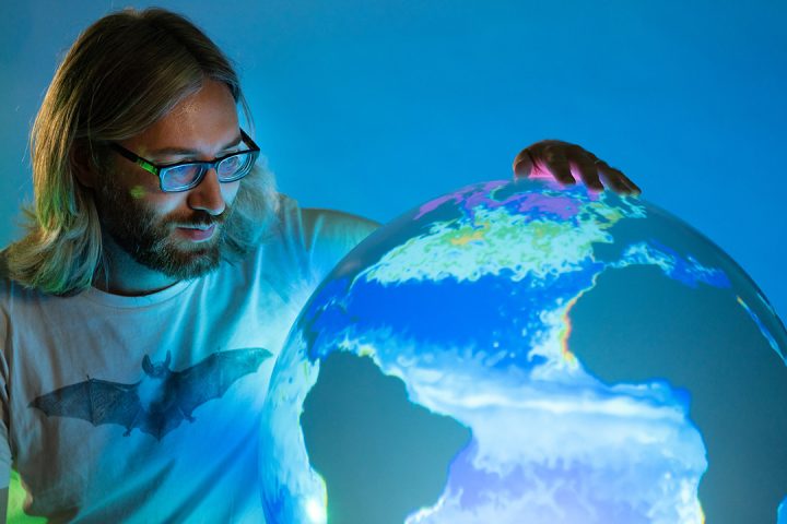 Person with long hair wearing glasses stands next to an illuminated globe, displaying a map of the world.