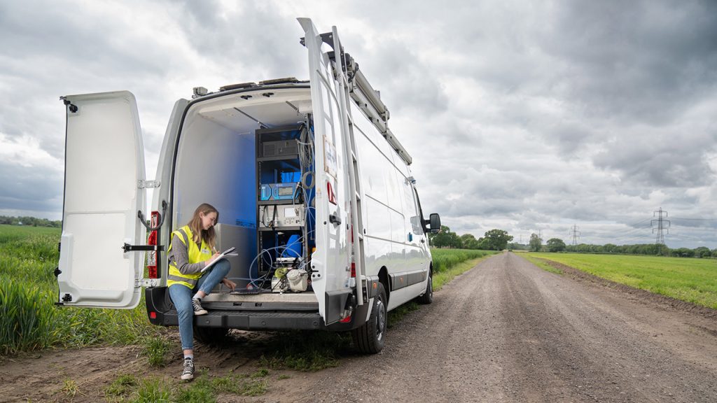 Person wearing high-vis jacket sits on the rear step on a large white van, holding a clipboard and pen. The van is filled with air pollution sensing equipment.