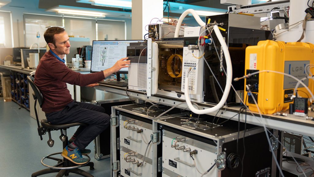 Person sits at a computer desk in a large laboratory space. The laboratory desk is filled with scientific equipment.