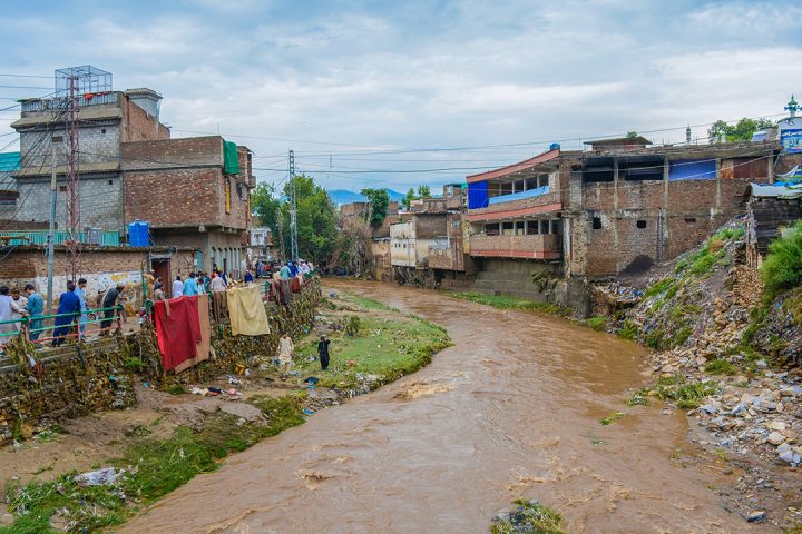 Muddy brown river water flows between brick buildings and piles of rubble and rubbish