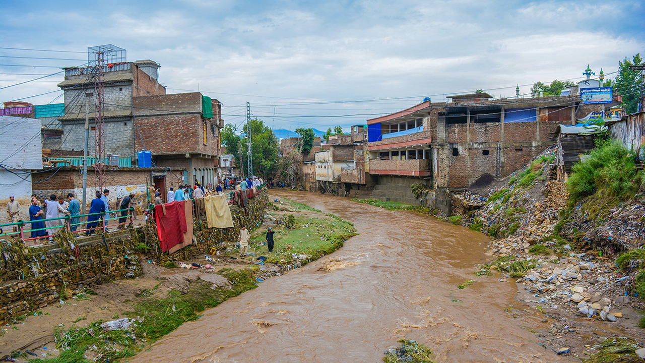 Muddy brown river water flows between brick buildings and piles of rubble and rubbish