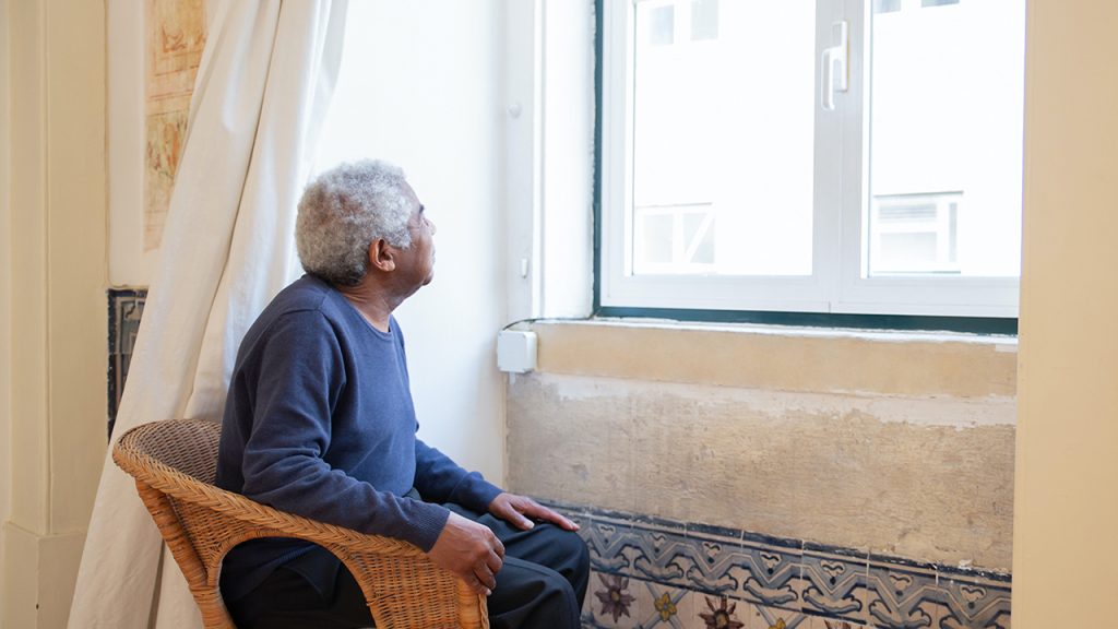 A old person of colour sat in a light brown wicker chair inside a building, looking out of a closed window