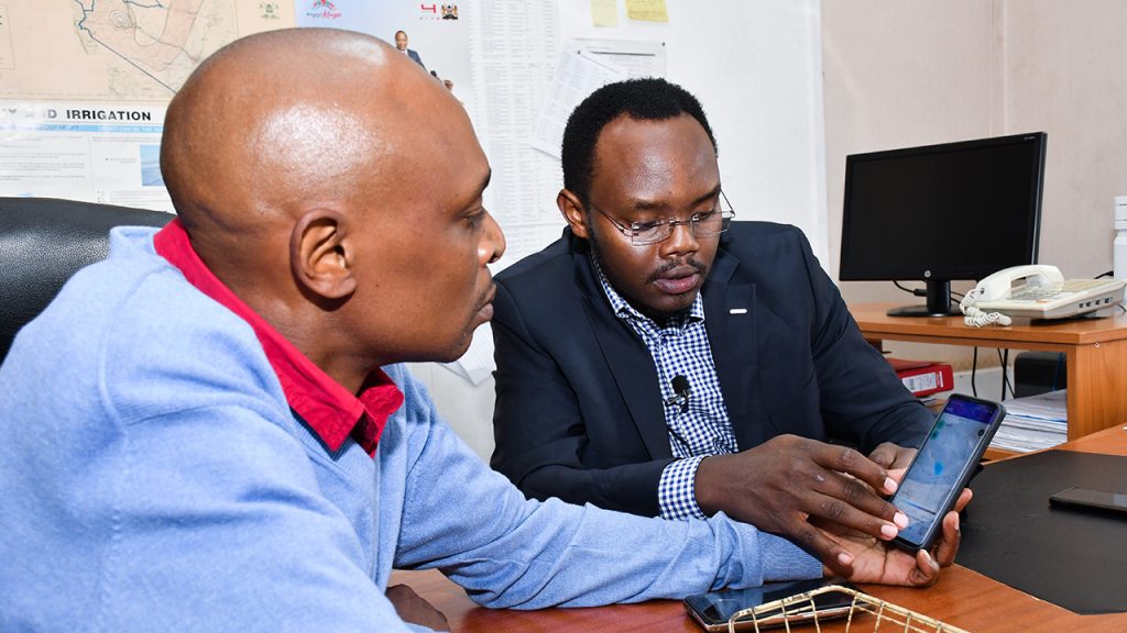 Two men sit at a desk, one demonstrates a mobile phone application which displays rainfall over Kenya