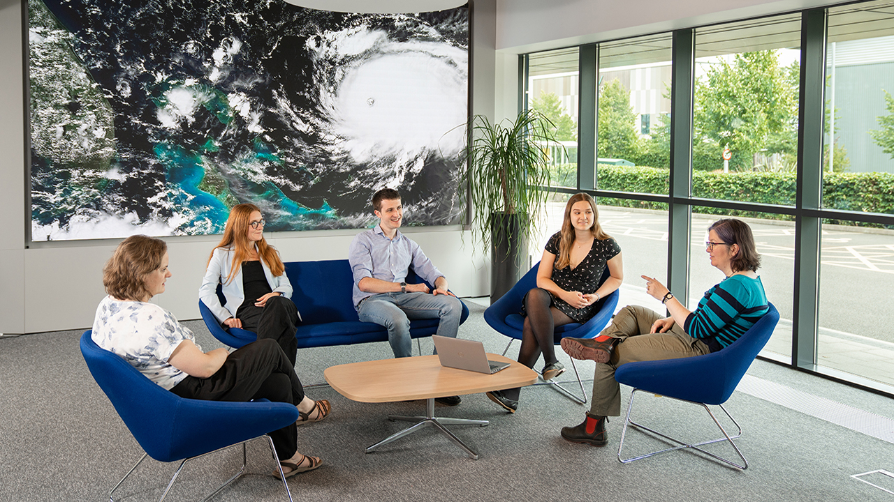 Five people sit on blue chairs around a small table, with a large picture in the background of a hurricane from a satellite