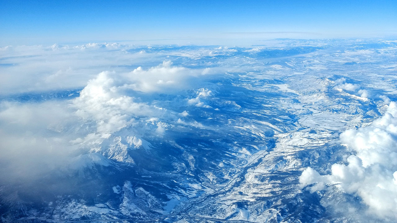 A photograph of a mountain range taken from above with clouds over them. The image is blue and white in colour.