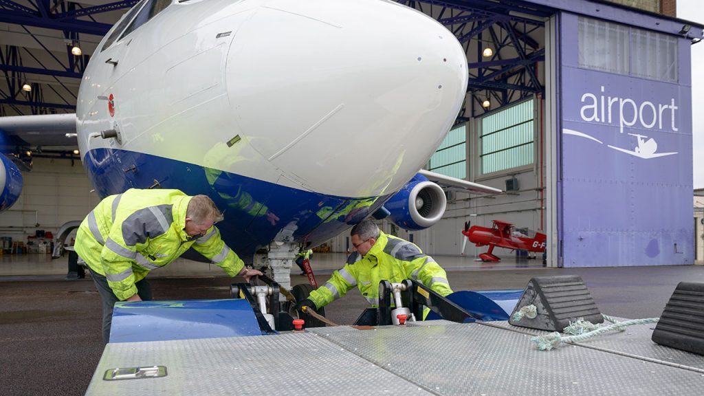 Two people wearing high-visibility jackets crouch underneath a large research aircraft front wheel, carrying out maintenance.