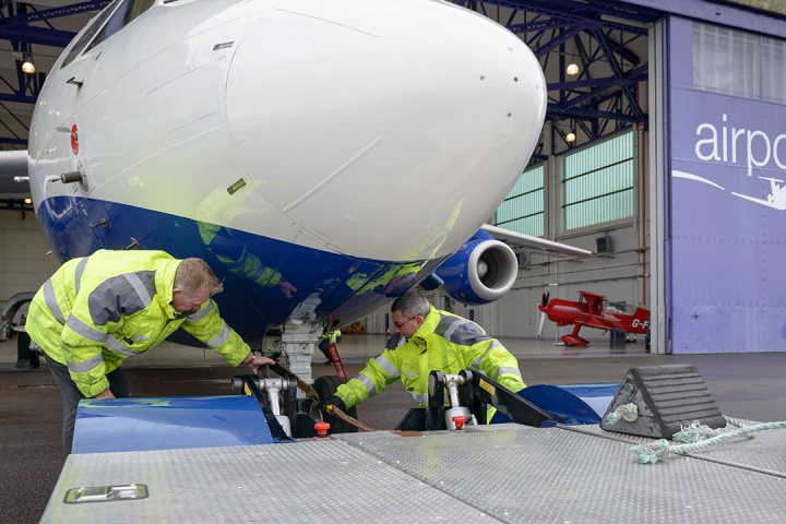 Two people wearing high-visibility jackets crouch underneath a large research aircraft front wheel, carrying out maintenance.