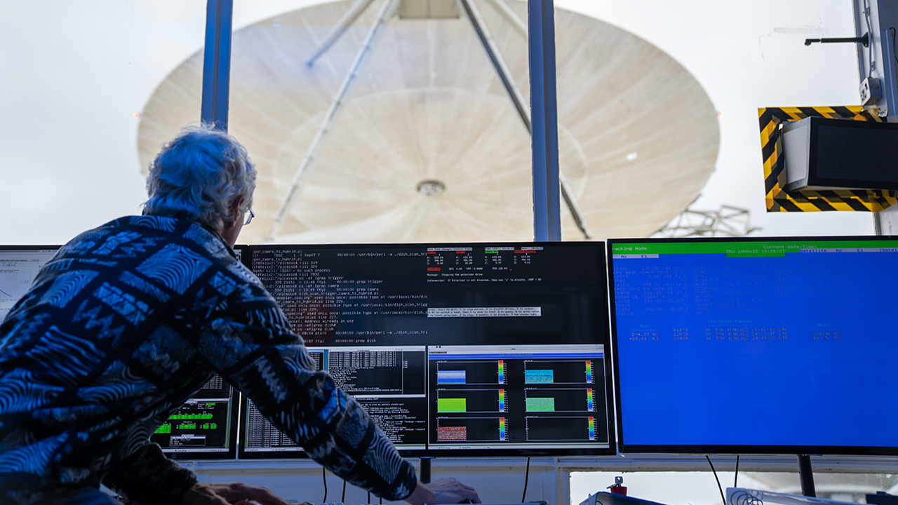 Person wearing patterned jumper leans over large computer desk with multiple screens. In front of him, through the window, he remotely controls a large, white weather radar dish.