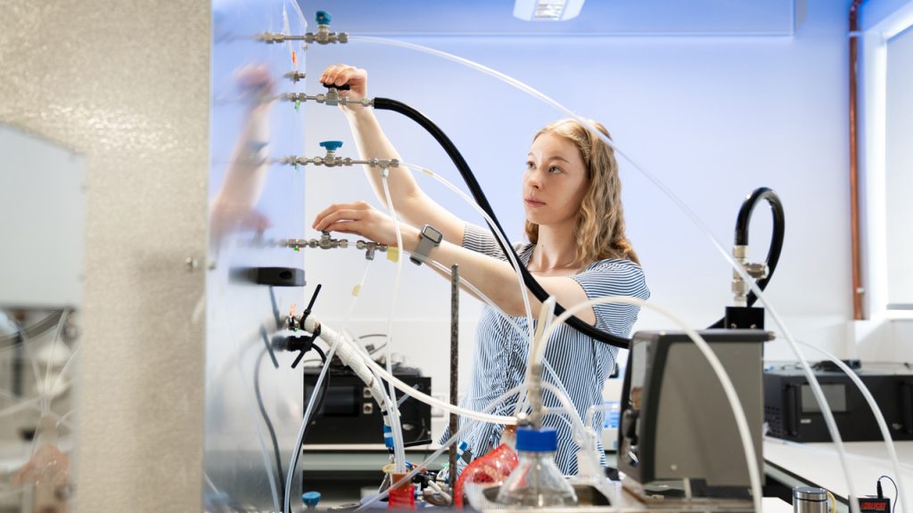 Person with long hair stands in a laboratory, wearing blue top and blue gloves. They reach upwards to adjust taps on the side of a large metal gas chamber.