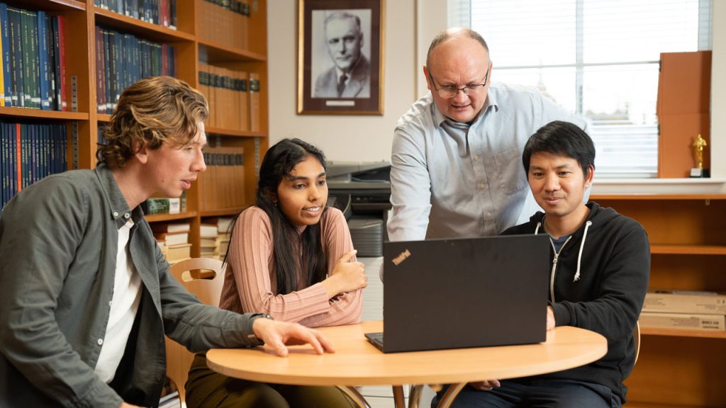 Three students sit round a wooden circular table, looking at a laptop. A man leans over them, pointing to something on the laptop. There are shelves of books on the left hand side.