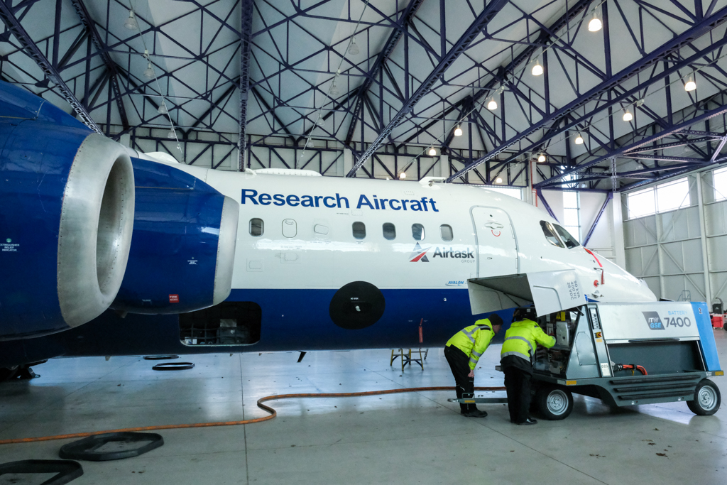 Two people wearing hi vis jackets are bent over looking into a power unti that is connected to a blu and white research aircraft inside a hangar