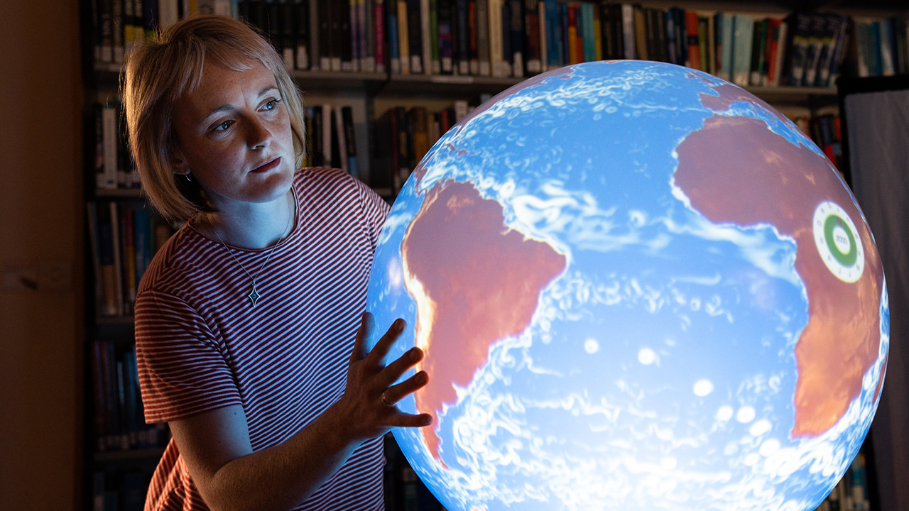 Person with shoulder-length hair and a striped shirt looks closely at a large illuminated globe, in a dark room. They touch the globe with both hands.