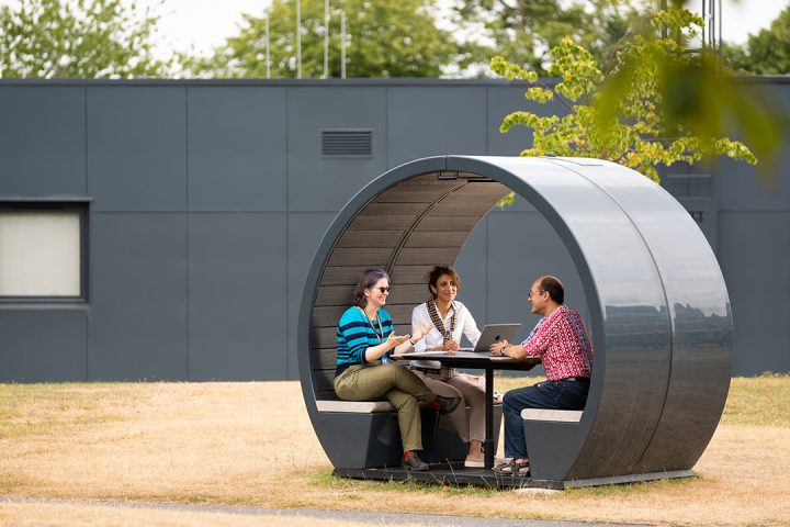 Three people sit around a small table in a grey cylinder shaped shelter in a leafy green outdoor space.