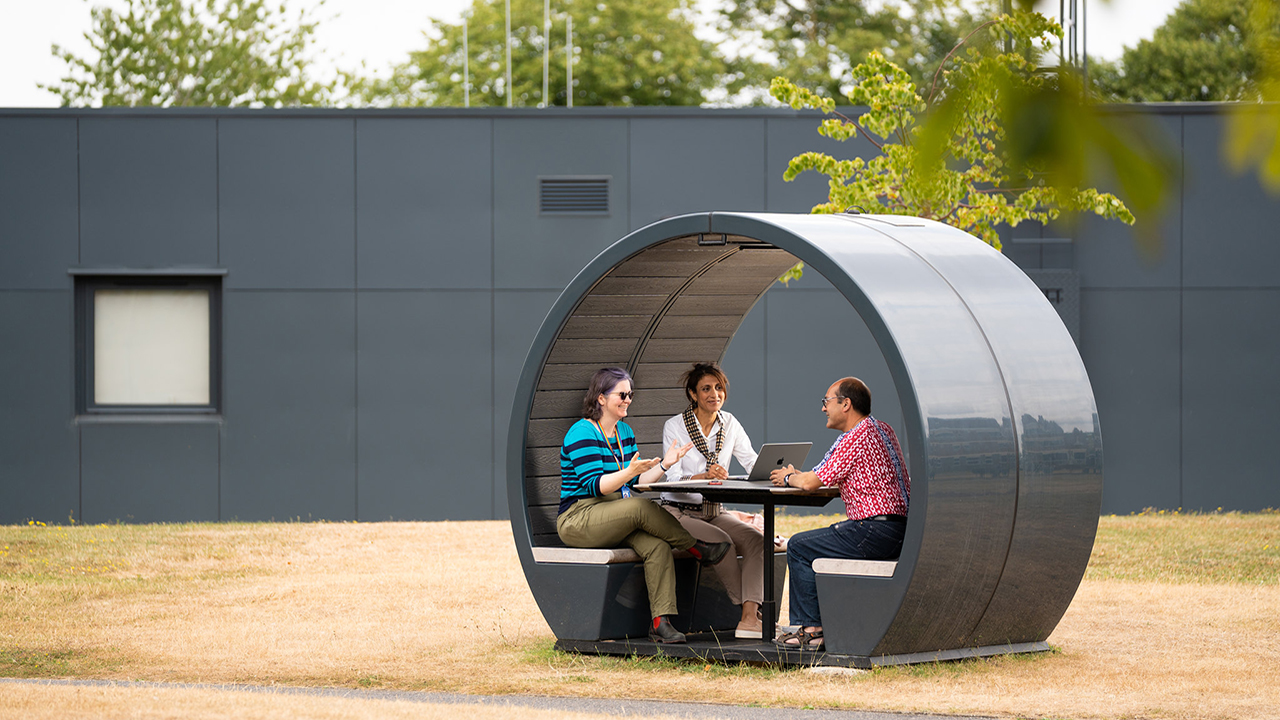 Three people sit around a small table in a grey cylinder shaped shelter in a leafy green outdoor space.