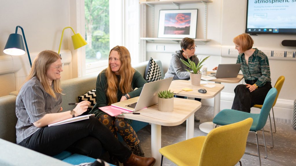 Four women sit around tables with laptops on in a comfy office space.