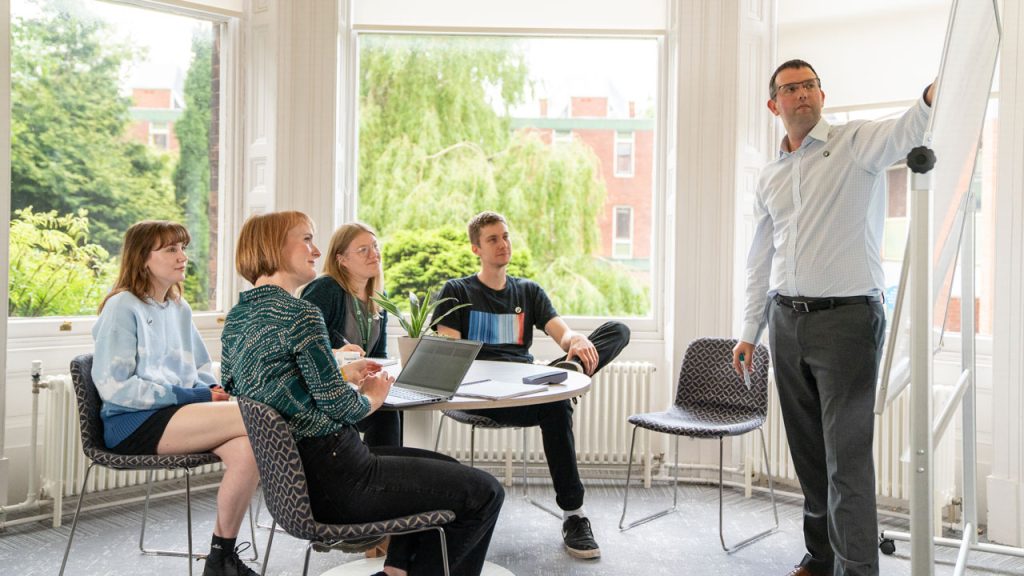 Four people sit around a circular table looking right towards a person standing pointing to a whiteboard. They are in front of sunny windows with trees in the background.