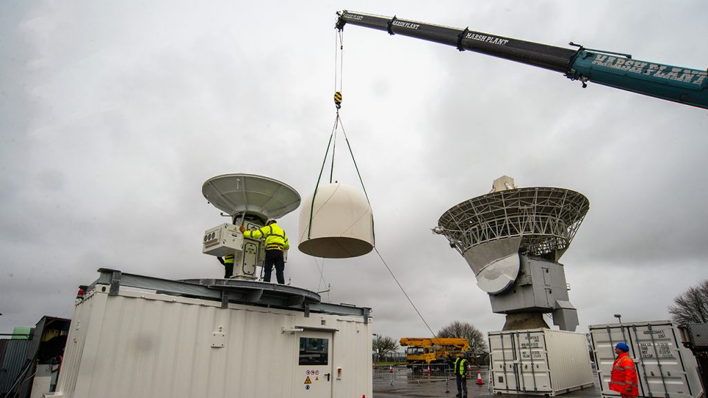 A crane lifts a white dome into the air, beside two radars, 3 shipping containers, and 4 people wearing hi vis jackets