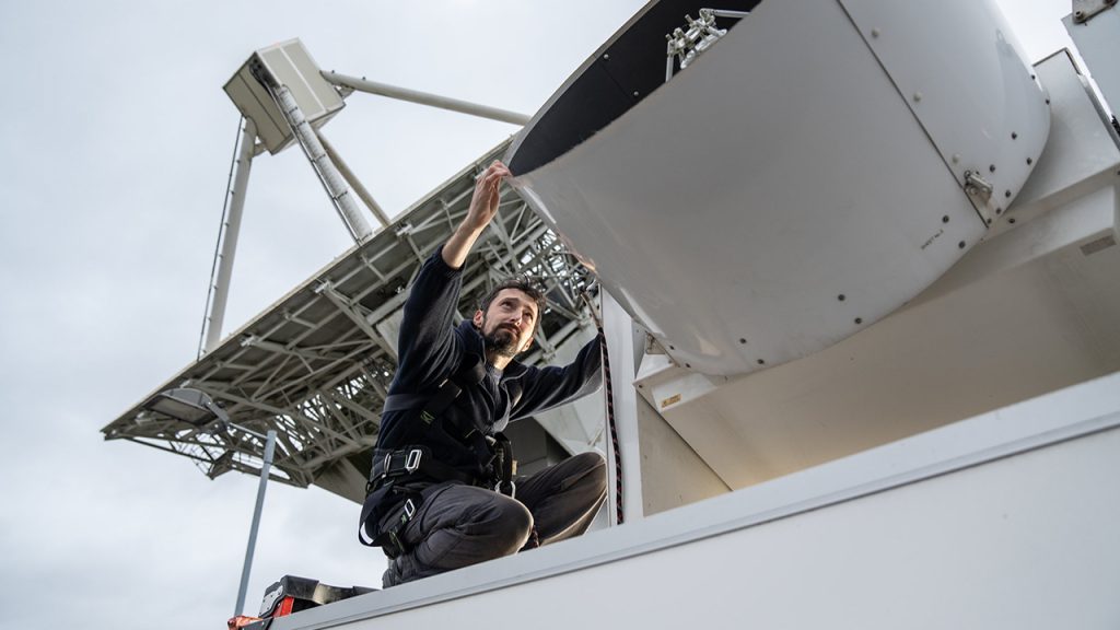 Person wearing blue overalls crouches next to a small cylindrical radar dish pointing skywards.