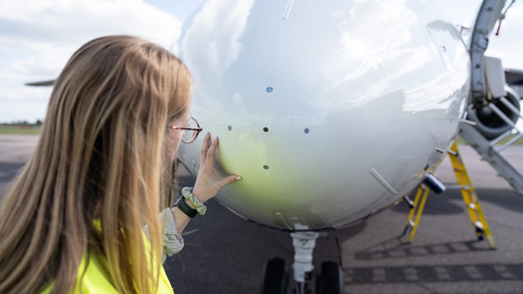 Person with shoulder length blonde hair wearing a hi vis jacket and glasses. They are looking at 4 holes that feed into scientific sensors inside the nose of a large white aircraft.