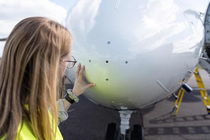 Person with shoulder length blonde hair wearing a hi vis jacket and glasses. They are looking at 4 holes that feed into scientific sensors inside the nose of a large white aircraft.