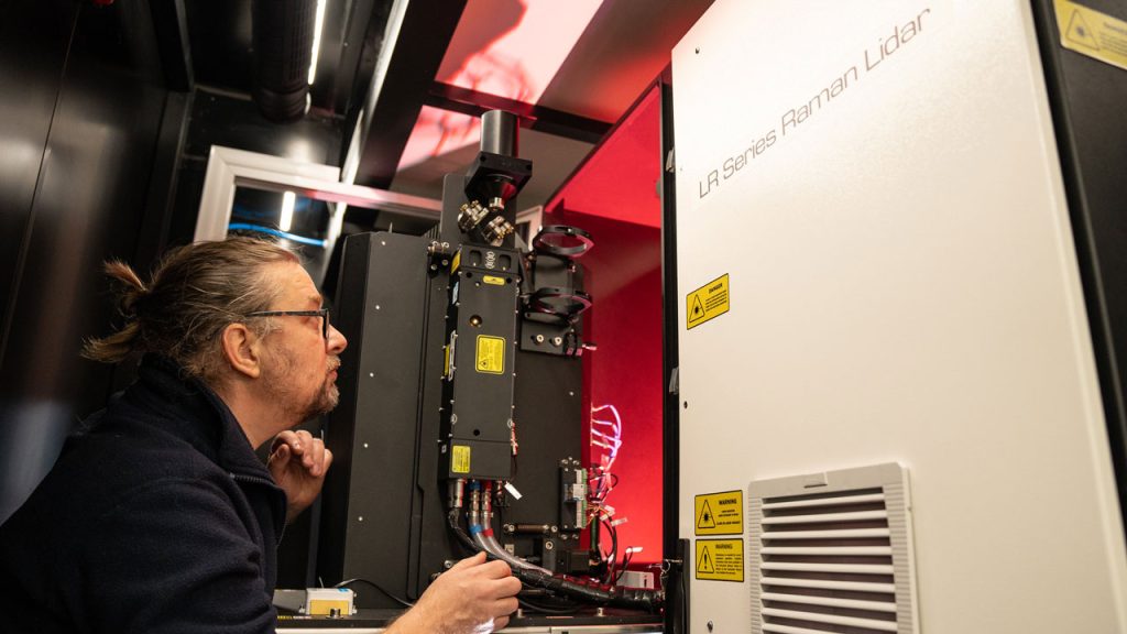 man crouches down in bottom left of the image, looking up at a lidar instrument.