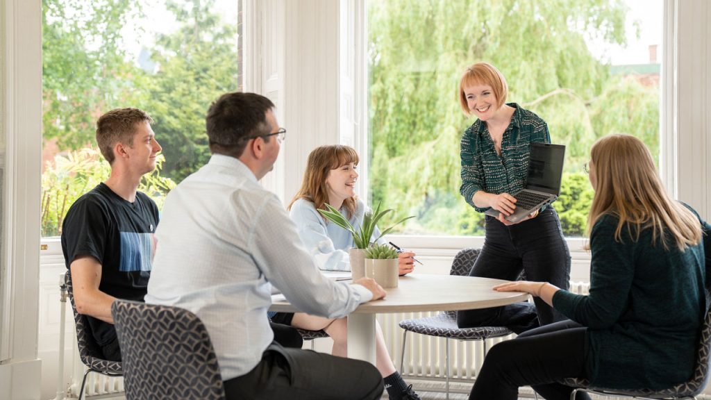 Four people sit around a circular table with one person knelt on a chair, holding a laptop. Large windows are behind them all with a view of green trees outside/