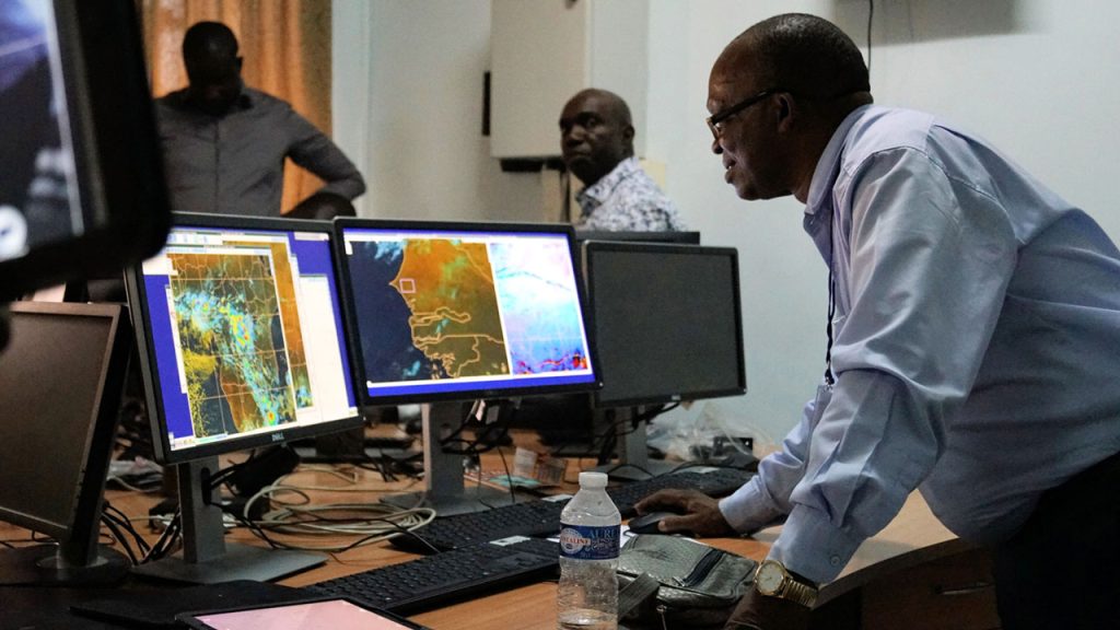 A man looks towards two computer screens with weather forecasting information on them. Two men stand in the background.