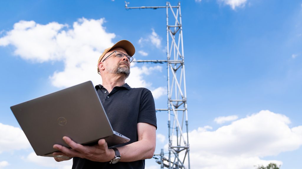 Person wearing glasses and dark blue shirt stands outside, looking towards the sky. They stand with a silver laptop in their hands.