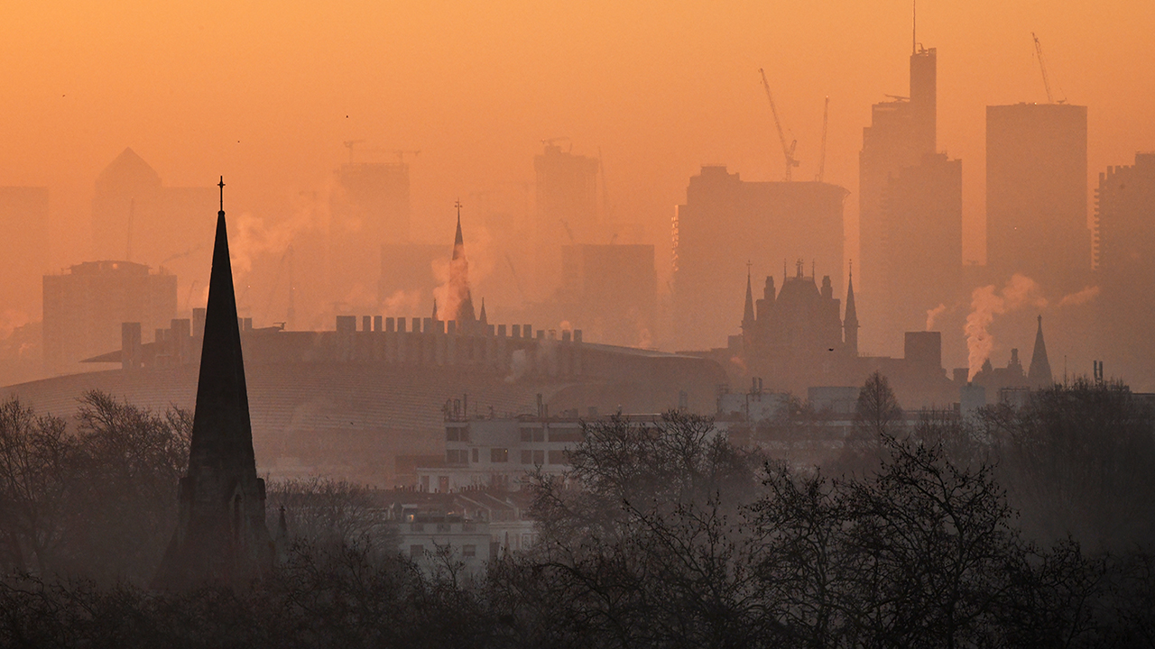 The view over the London cityscape at dawn from Primrose Hill in the north of the London. The sky is orange and hazy, and there are puffs of air rising from building, and tree silhouettes in the foreground.