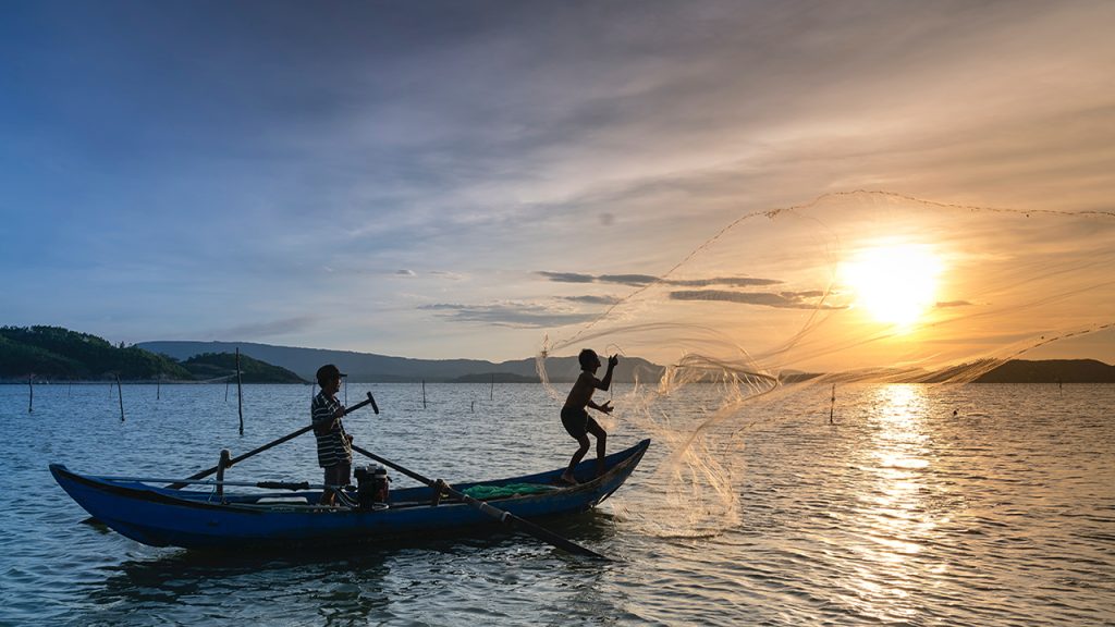Two people on a boat casting a net onto the water with the sun low in the sky.