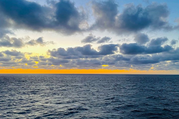 Blue ocean with clouds in the sky and sun setting over the water.