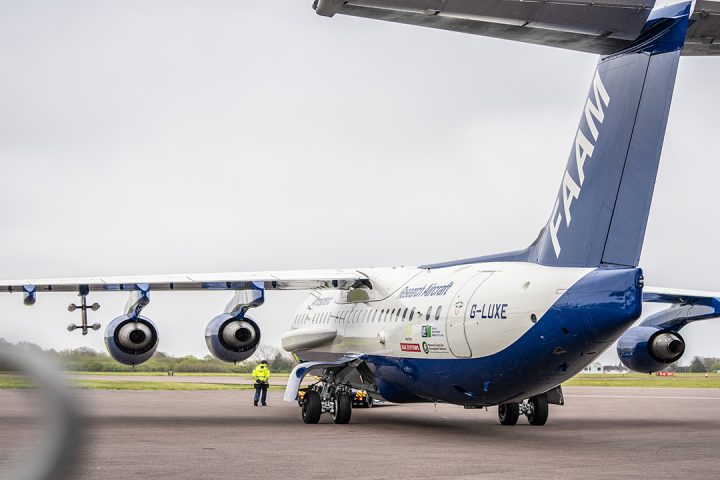 Blue and white research aircraft being towed across apron with person wearing high visibility jacket walking next to it.