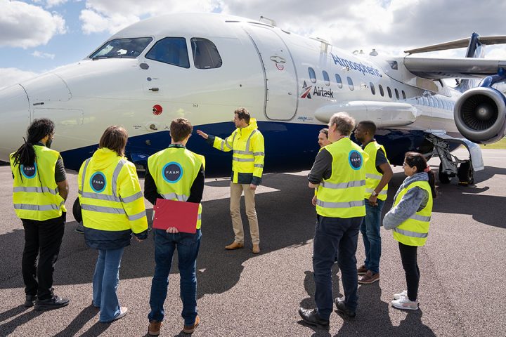 Eight people wearing high visibility jackets look at blue and white research aircraft. One person is pointing towards the aircraft. Blue sky and clouds in background.