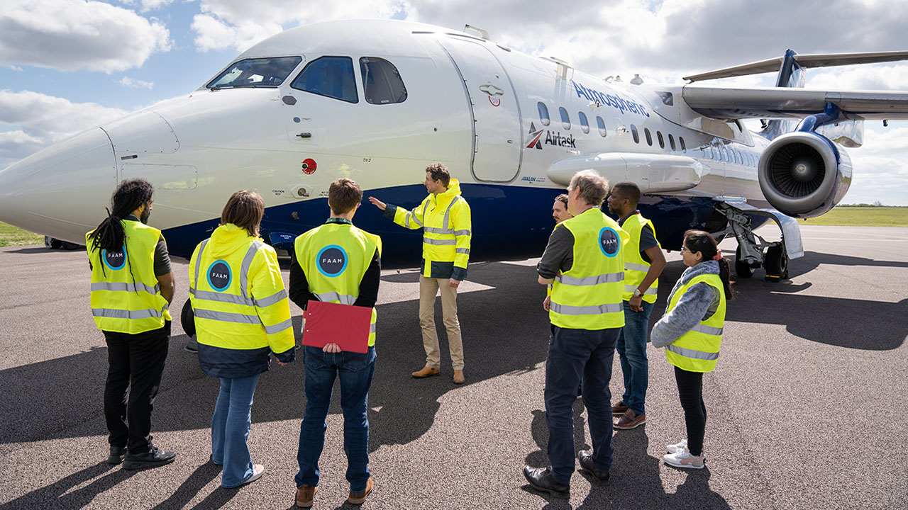 Eight people wearing high visibility jackets look at blue and white research aircraft. One person is pointing towards the aircraft. Blue sky and clouds in background.