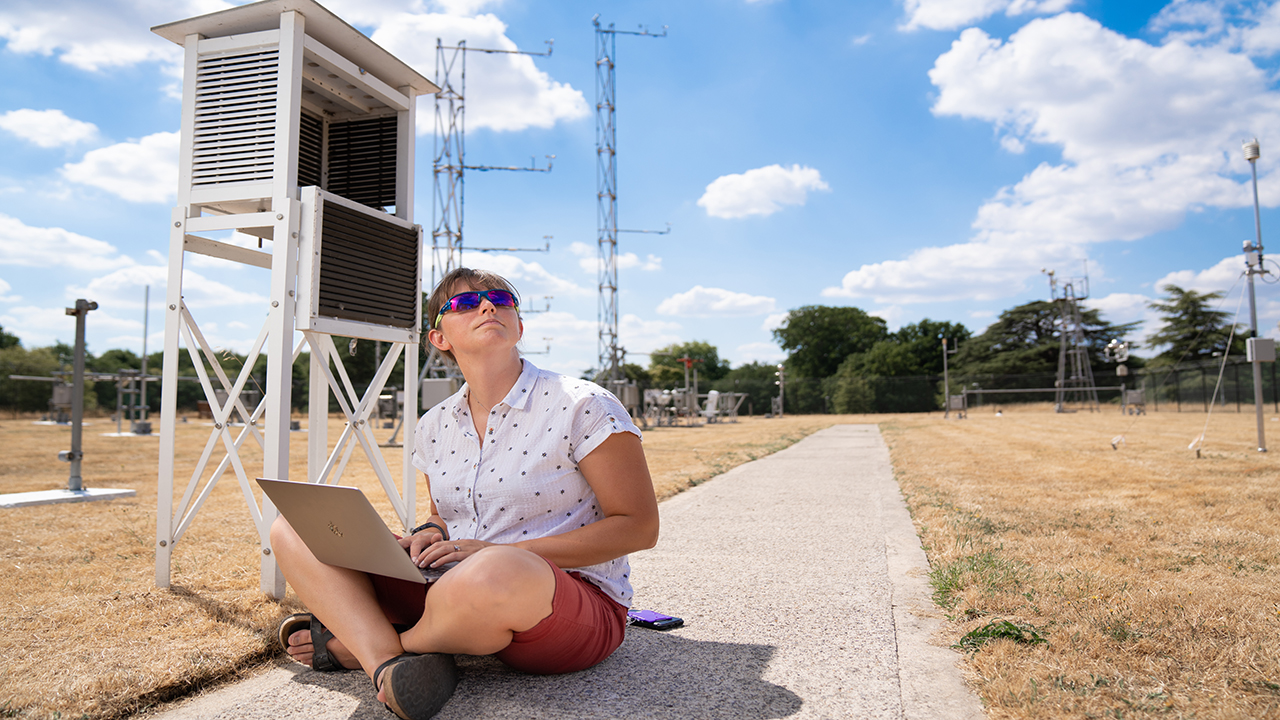 Person wearing sunglasses sits outside in a field full of scientific antenna. They hold a laptop and look up towards the blue sky above.