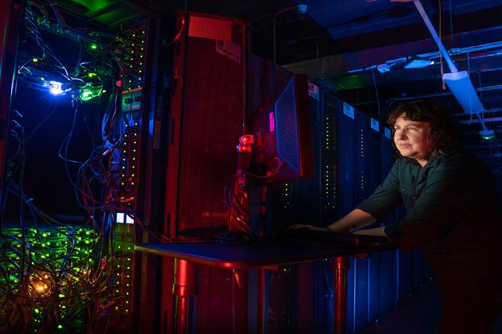 Person with dark curly hair, wearing a green shirt stands at a mobile computer desk in a dark room full of computer equipment.