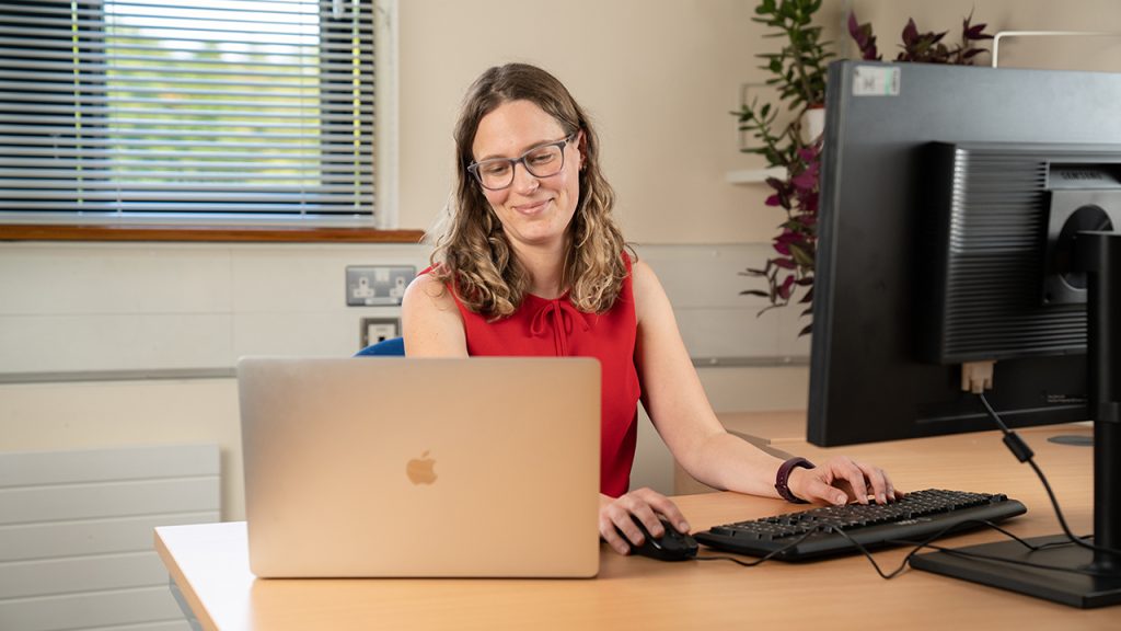 Person wearing glasses and red top sits at a computer desk inside, they look towards a silver laptop on the table.