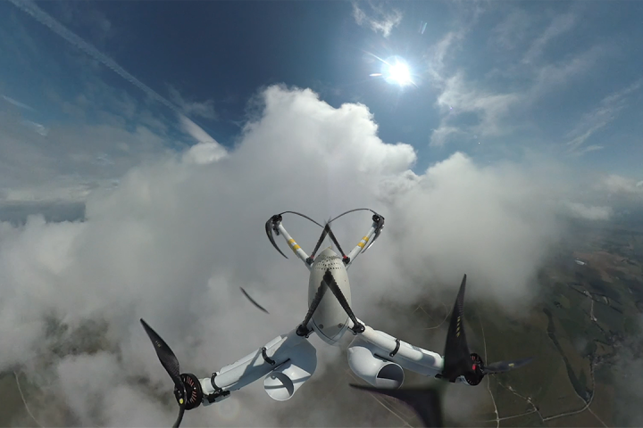 Small weather sensing drone flying upwards through white clouds and blue skies with green land below.