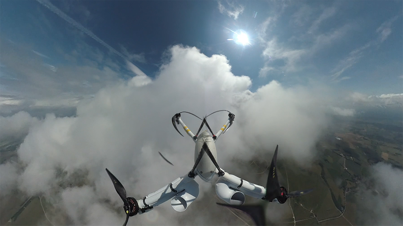 Small weather sensing drone flying upwards through white clouds and blue skies with green land below.