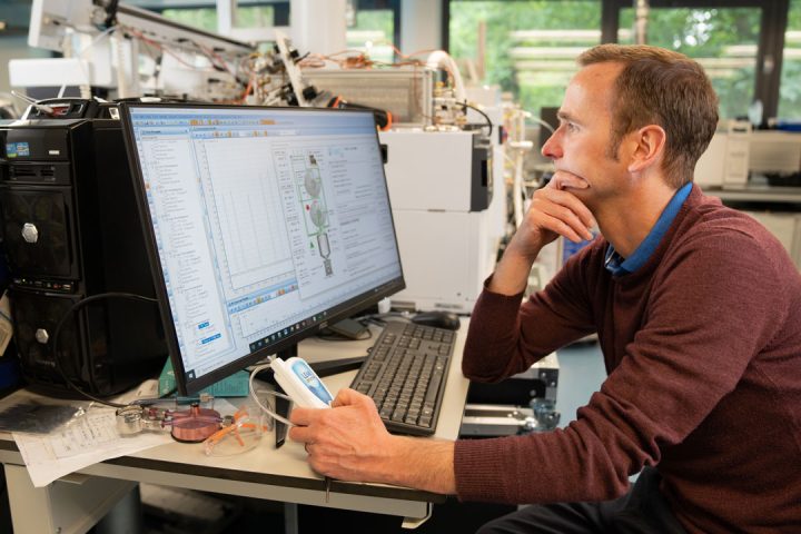 A male scientist looks at computer screens with scientific information on them. They are sat in a laboratory.