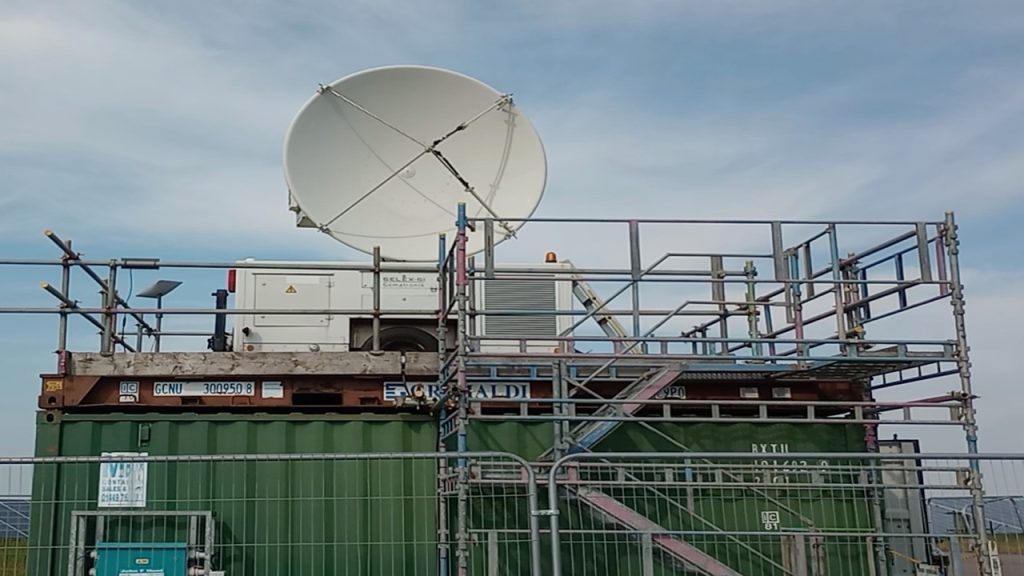 Large white radar on top of green metal container surrounded by metal scaffolding. Blue sky with clouds in background.
