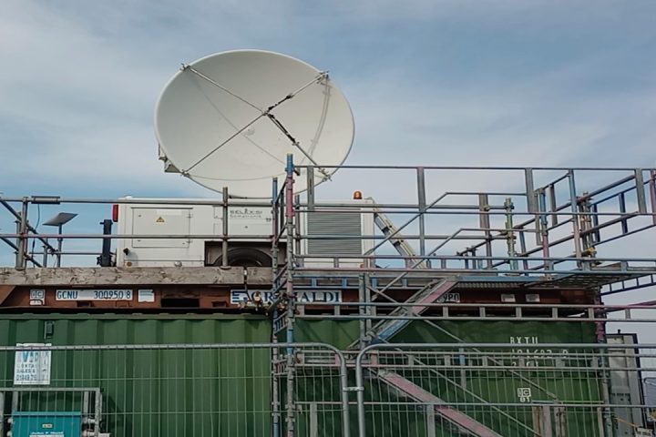 Large white radar on top of green metal container surrounded by metal scaffolding. Blue sky with clouds in background.