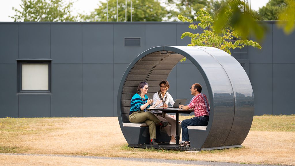 Three people sat inside a grey dome, surrounded by dry grass and green trees