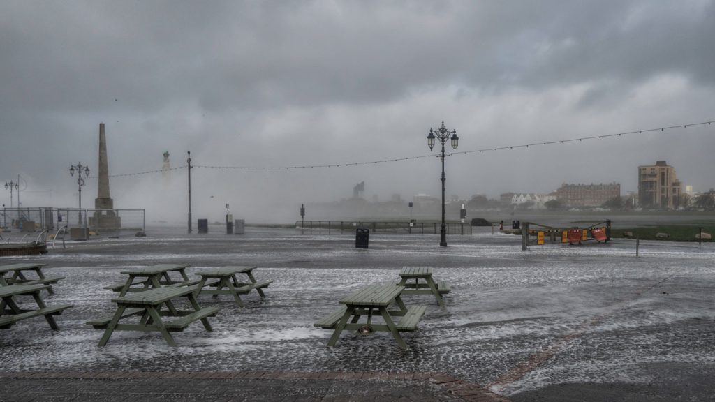 Seafront during storm. Green picnic benches surrounded by water. Grey clouds and seafoam in background.