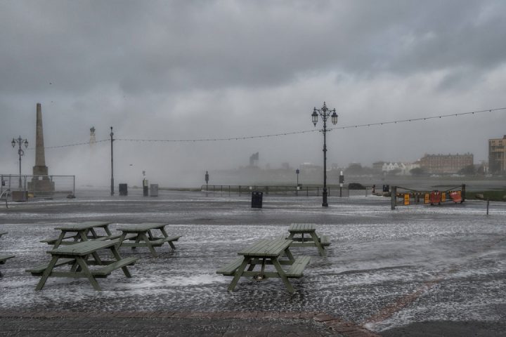 Seafront during storm. Green picnic benches surrounded by water. Grey clouds and seafoam in background.